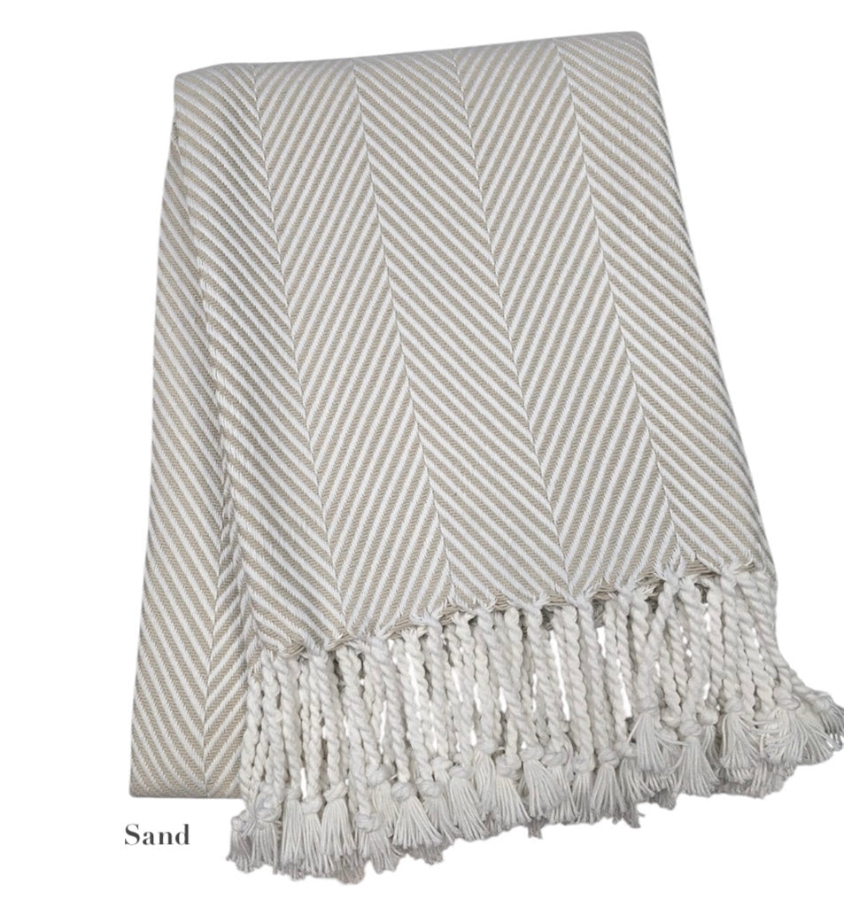 The Nantucket Herringbone Throw Collection by A Soft Idea in Sand features a beige and white cotton throw with hand-knotted fringe, folded neatly.