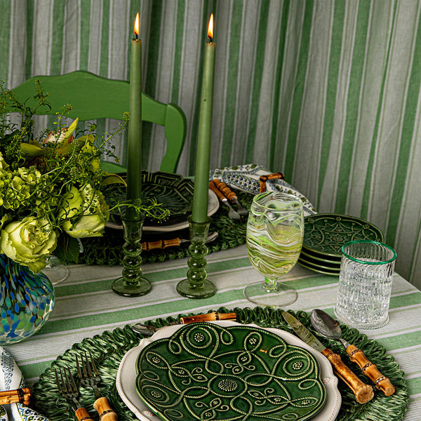 A table set with green patterned plates, bamboo-handled cutlery, a Juliska Provence Candlestick Set, two tall green candles, a vibrant green glass, a clear tumbler, flowers, and a striped green tablecloth.