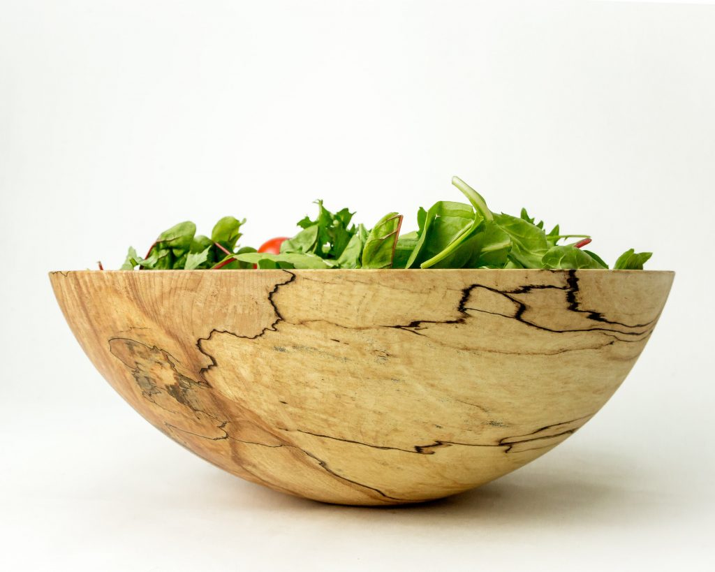 A mixed green salad is served in a Peterman's Spalted Maple Serving Bowl, showcasing its unique grain pattern against a plain white background.