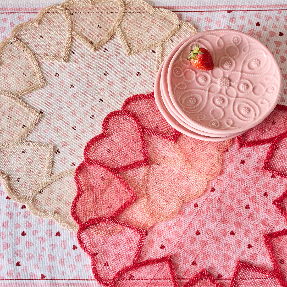 Juliska's Heart Placemat Collection features pink and red hand-woven abaca placemats, paired with pink plates and a strawberry accent on a heart-pattern tablecloth—an elegant tablescape perfect for any celebration.