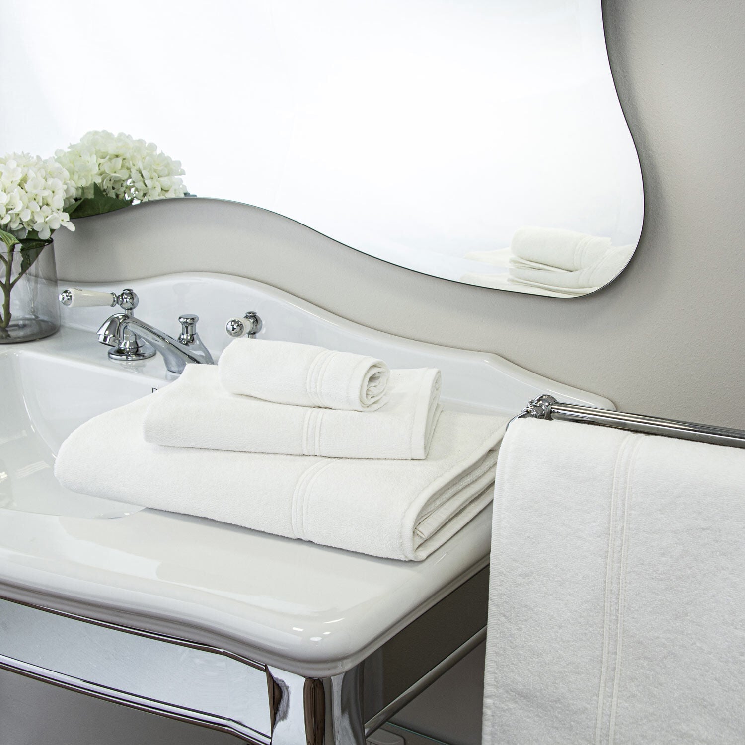 A stack of Frette's Classic White/White Bath Collection towels sits on a bathroom sink beside a white floral arrangement, with an extra towel hanging nearby, complementing the elegant decor.