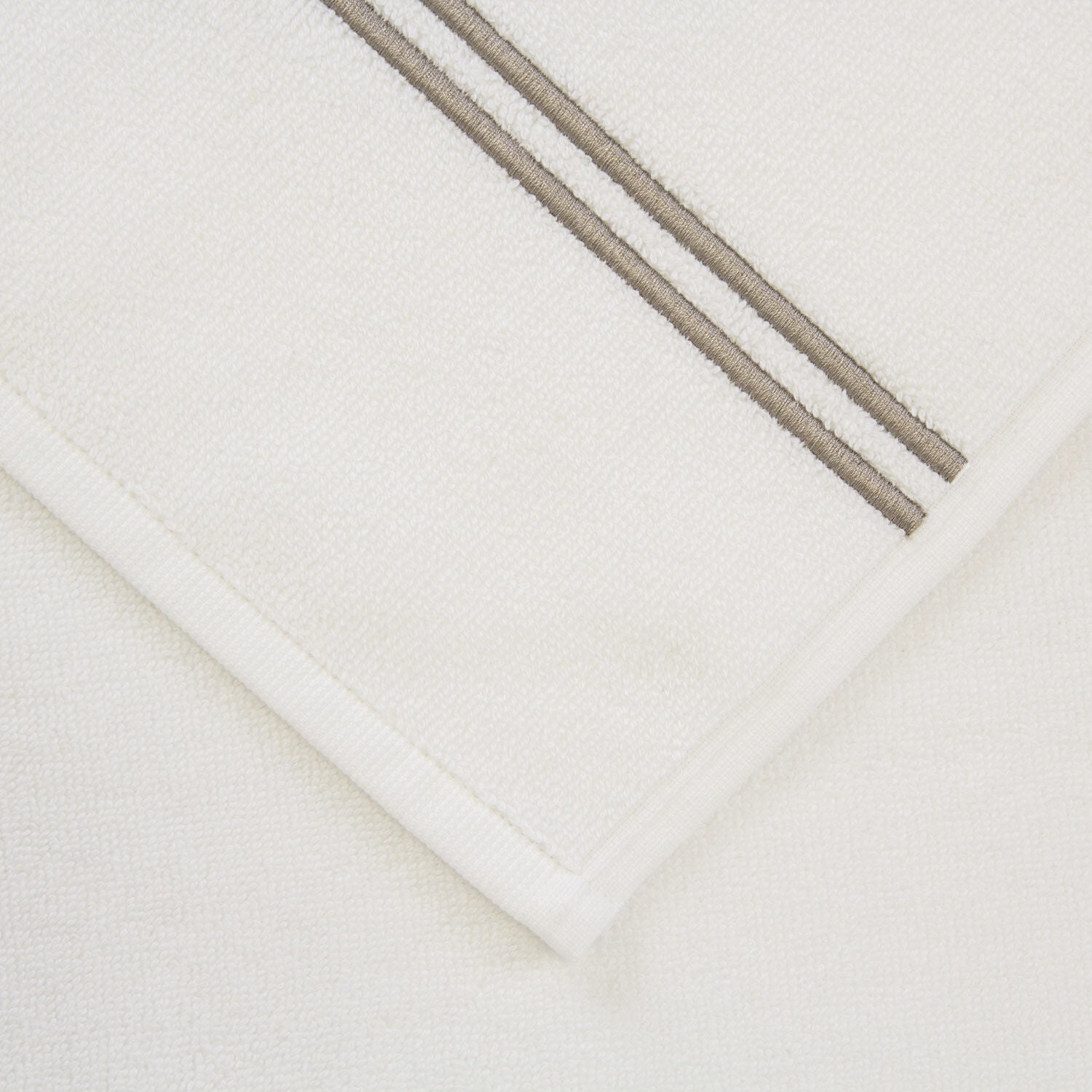 A folded white towel from Frette’s Classic White/Khaki Bath Collection, crafted in soft cotton terry with two parallel khaki stripes, adds a stylish touch to your bathroom.