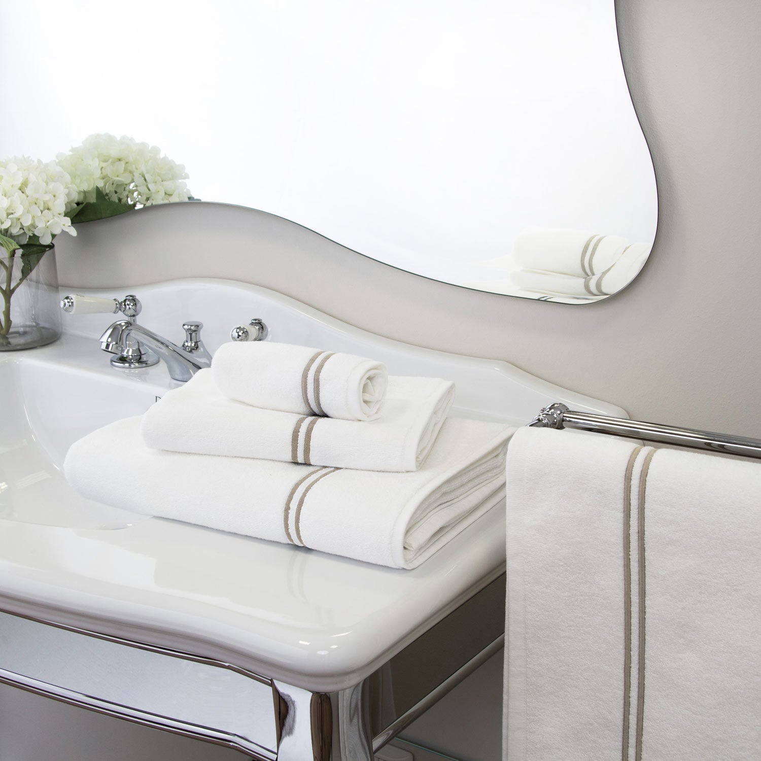Frette’s Classic White/Khaki Bath Collection, crafted from soft cotton terry, is neatly folded on the sink, adding elegance and complementing the white flowers and matching towel by the mirror in this stylish bathroom.