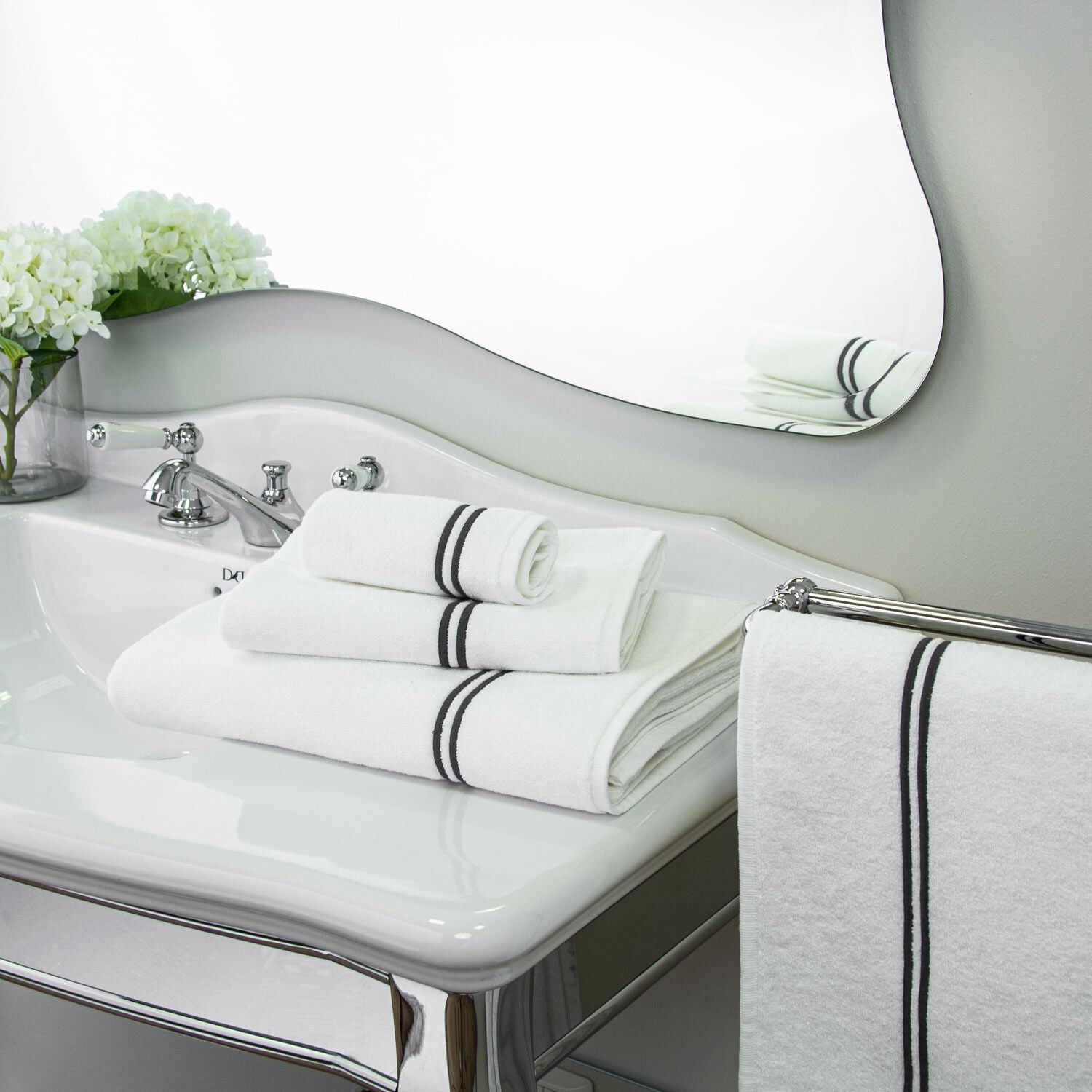 The Classic White/Grey Bath Collection by Frette features neatly folded towels stacked on a bathroom sink, with a matching towel hanging on the side and white flowers in the background for an elevated bathroom experience.