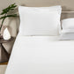 A neatly made bed features the Frette Classic White/White Bedding Collection, crafted from 100% cotton, with pillows beside a nightstand displaying a vase with a leaf and a white decorative object.