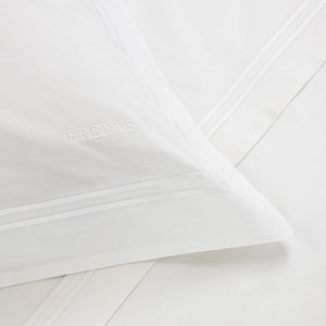 A close-up of the Frette Classic White/White Bedding Collection showcases a white pillowcase with an embroidered logo and matching 100% cotton bedding, featuring subtle decorative borders for a luxurious touch.