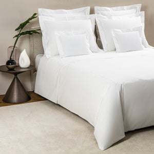 A neatly made bed features Frette’s Classic White/White Bedding Collection with multiple pillows, beside a side table displaying a plant in a glass vase and a white decorative object.