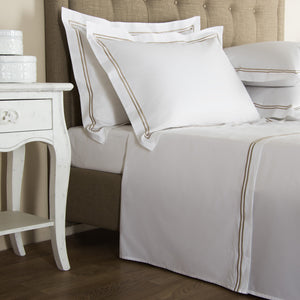 A neatly made bed features the Frette Classic White/Khaki Bedding Collection, its white linens with beige trim beside a white nightstand with decorative boxes, all set on a wooden floor.
