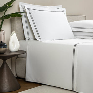 A neatly made bed with white linens featuring black trim from the Frette Classic Bedding Collection, White/Grey, two pillows, and a bedside table with a vase, decorative bowl, and plant. The luxurious sheet set from Frette is 100% cotton.