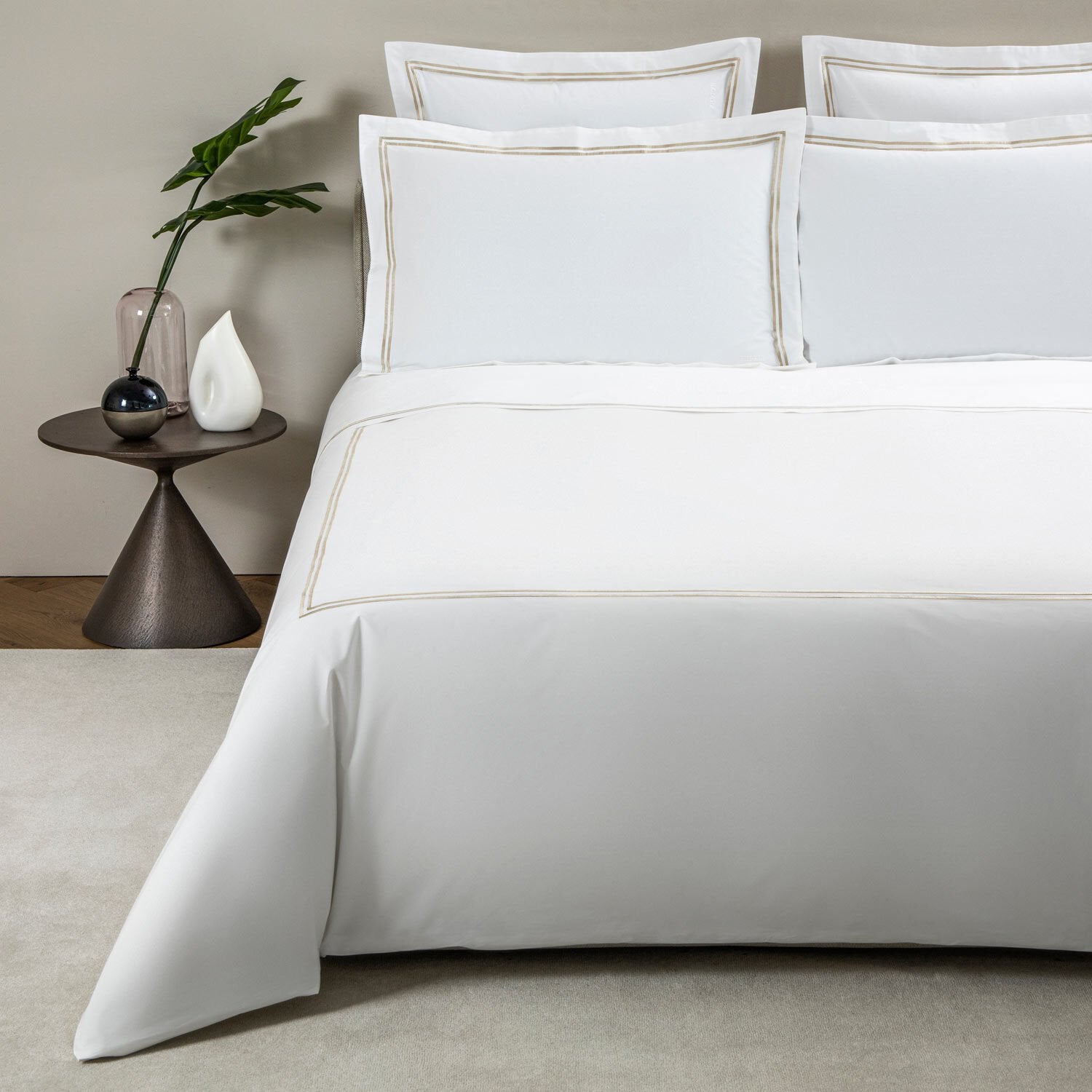 A bed made with Frette’s Classic White/Khaki Bedding Collection is paired with matching pillows and a round side table displaying a vase with a green plant, glass orb, and white decorative object from the classic collection.