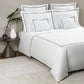 A bed dressed in Frette’s Classic White/Grey Bedding Collection features luxurious 100% cotton sheets with grey trim, coordinating pillows, and is styled beside a table with a plant, glass vase, and decorative white object.