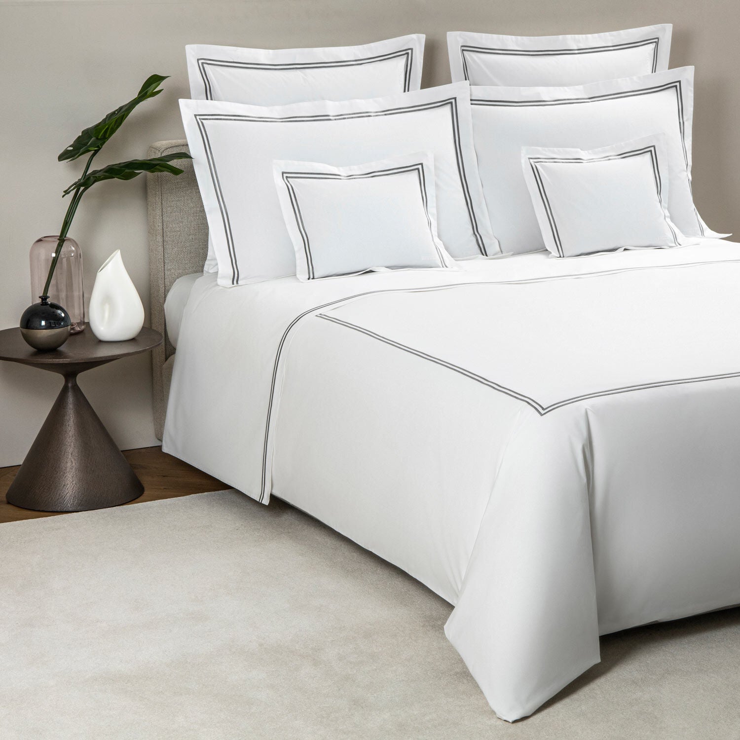 A bed dressed in Frette’s Classic White/Grey Bedding Collection features luxurious 100% cotton sheets with grey trim, coordinating pillows, and is styled beside a table with a plant, glass vase, and decorative white object.