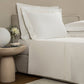 A neatly made bed with Frette's Triplo Bourdon White/Milk Bedding Collection, featuring white poplin sheets and three-line embroidery, stands next to a round bedside table with a glass vase of flowers, white lamp, and cup and saucer.