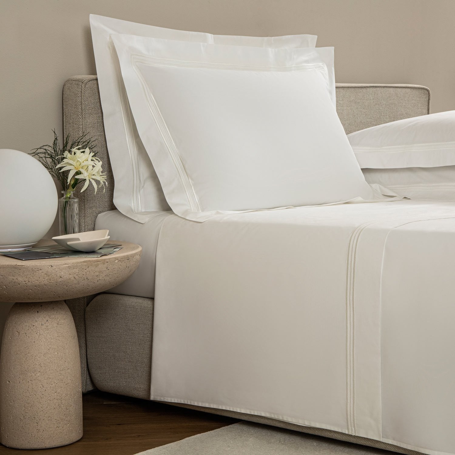 A neatly made bed with Frette's Triplo Bourdon White/Milk Bedding Collection, featuring white poplin sheets and three-line embroidery, stands next to a round bedside table with a glass vase of flowers, white lamp, and cup and saucer.