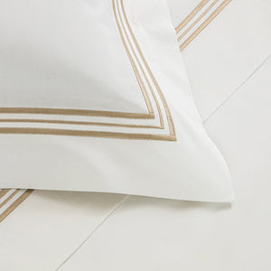 A close-up of a white pillow with beige embroidered borders from Frette's Triplo Bourdon White/Beige Bedding Collection, crafted in cotton poplin and shown on coordinating bedding. Made in Italy.
