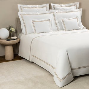 A neatly made bed with Frette’s Triplo Bourdon White/Beige Bedding Collection—featuring white cotton poplin and tan borders—plus multiple pillows, a round side table with a lamp, book, and small vase. Luxuriously crafted in Italy.