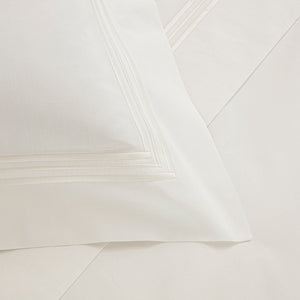 A close-up of a neatly made bed with a white pillow from Frette's Triplo Bourdon White/Milk Bedding Collection, featuring elegant three-line embroidery near the border, resting on matching cotton poplin white sheets.
