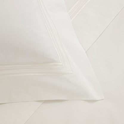 A close-up of a neatly made bed with a white pillow from Frette's Triplo Bourdon White/Milk Bedding Collection, featuring elegant three-line embroidery near the border, resting on matching cotton poplin white sheets.