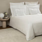 A neatly made bed with Frette’s Triplo Bourdon White/Milk Bedding Collection, featuring elegant three-line embroidery, is accented by multiple pillows and a round stone nightstand topped with a spherical lamp and a small flower vase.