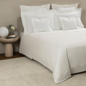 A neatly made bed with Frette’s Triplo Bourdon White/Milk Bedding Collection, featuring elegant three-line embroidery, is accented by multiple pillows and a round stone nightstand topped with a spherical lamp and a small flower vase.