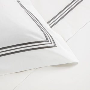 A close-up of a white pillowcase from Frette's Triplo Bourdon White/Grey Bedding Collection, detailed with two dark grey embroidered stripes along the edge, resting on a coordinating white cotton poplin sheet.
