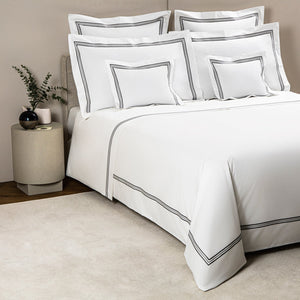 A neatly made bed with Frette's Triplo Bourdon White/Grey Bedding Collection, featuring grey double-line borders, matching pillows, and a round nightstand adorned with a vase and decorative accents.