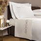 The Links Embroidery Bedding Collection by Frette features white cotton sateen sheets and pillowcases with elegant chain embroidery, shown on a dark bed beside a marble-topped nightstand decorated with accents and a vase of dried plants.