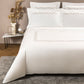 A bed dressed in Frette’s Links Embroidery Bedding Collection, showcasing white cotton sateen sheets with an elegant chain motif, is flanked by pillows and accompanied by a marble side table holding a vase, dried plants, and a lamp.