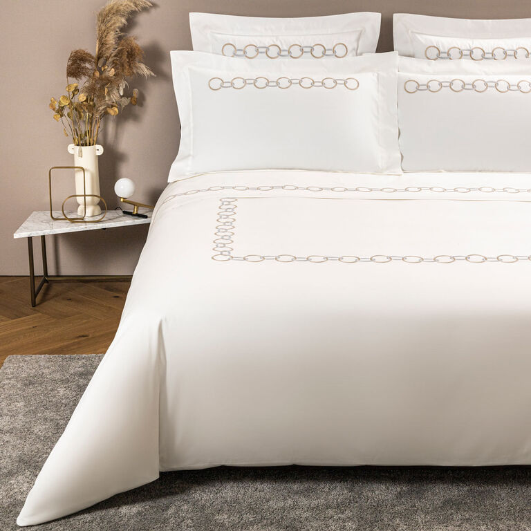 A bed dressed in Frette’s Links Embroidery Bedding Collection, showcasing white cotton sateen sheets with an elegant chain motif, is flanked by pillows and accompanied by a marble side table holding a vase, dried plants, and a lamp.