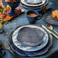 Pieces from the Juliska Le Panier Mystic Knot Denim Dinnerware and Serveware Collection—including a blue and white patterned plate, bowl, and mug—are arranged on a blue tablecloth with flowers, cutlery, and sliced bread in the background. A large charger from the collection completes the elegant tablescape.