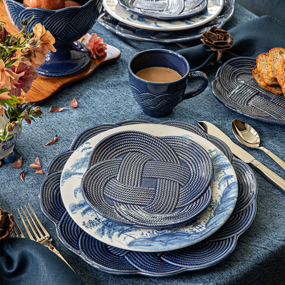 Pieces from the Juliska Le Panier Mystic Knot Denim Dinnerware and Serveware Collection—including a blue and white patterned plate, bowl, and mug—are arranged on a blue tablecloth with flowers, cutlery, and sliced bread in the background. A large charger from the collection completes the elegant tablescape.