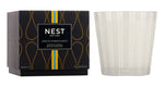 A Nest Amalfi Lemon & Mint Collection candle, inspired by the Amalfi Coast, is displayed beside its stylish black box featuring gold accents with yellow and blue stripes.