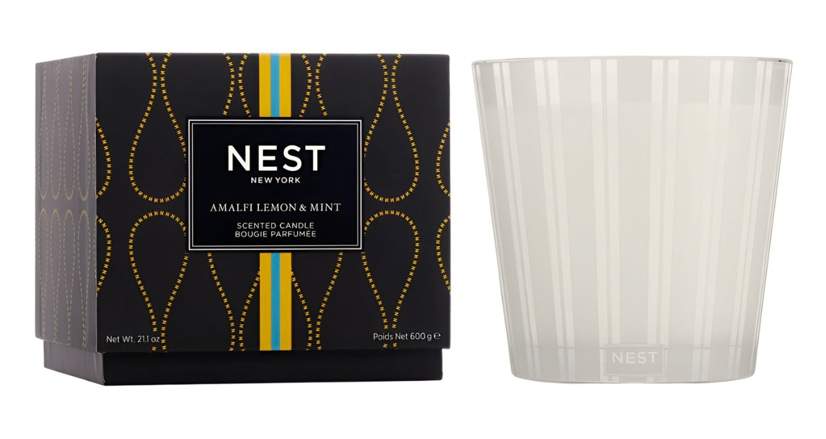 A Nest Amalfi Lemon & Mint Collection candle, inspired by the Amalfi Coast, is displayed beside its stylish black box featuring gold accents with yellow and blue stripes.