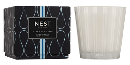 A Nest Ocean Mist & Sea Salt Candle from the Candle Collection sits in a striped glass holder beside its black and blue packaging, infusing the air with a refreshing ocean mist and sea salt aroma.
