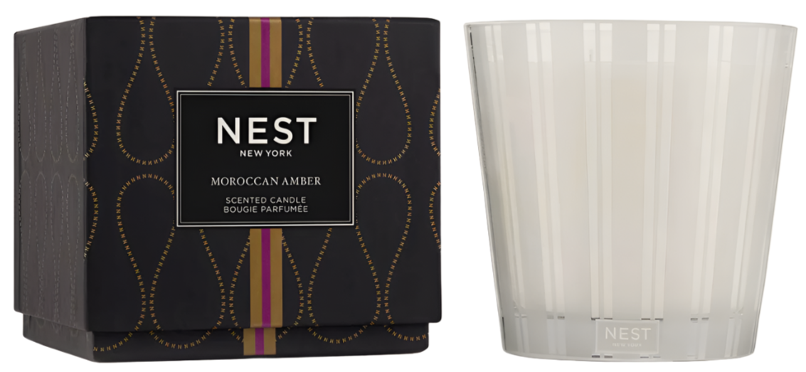A Nest Moroccan Amber Candle Collection box sits next to a clear glass candle, filling the air with warm notes of patchouli and bergamot.