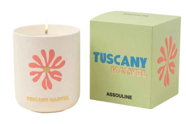 A lit candle in a white holder with a floral design casts a warm glow next to the green box labeled "Travel Candle Tuscany Marvel" by Assouline, releasing soothing amber notes that seamlessly blend into the enchanting fragrance of Tuscany.