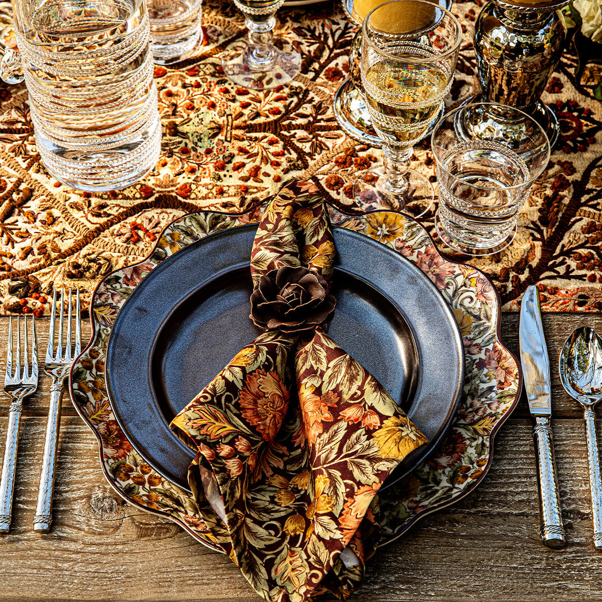 A formal table setting with floral-patterned plates featuring an elegant floral motif, the Willa Amber Napkin Set by Juliska, silver cutlery, glassware, and a decorative tablecloth.
