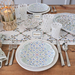 A wooden table set with patterned folkloric plates, silver cutlery, Villa Seville Chambray Place Cards by Juliska, white glasses, a candle in a wicker holder, and a floral table runner.