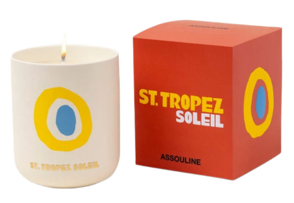 A colorful "Travel Candle St. Tropez Soleil" by Assouline, with ginger hints, sits beside an orange box displaying the same inscription.