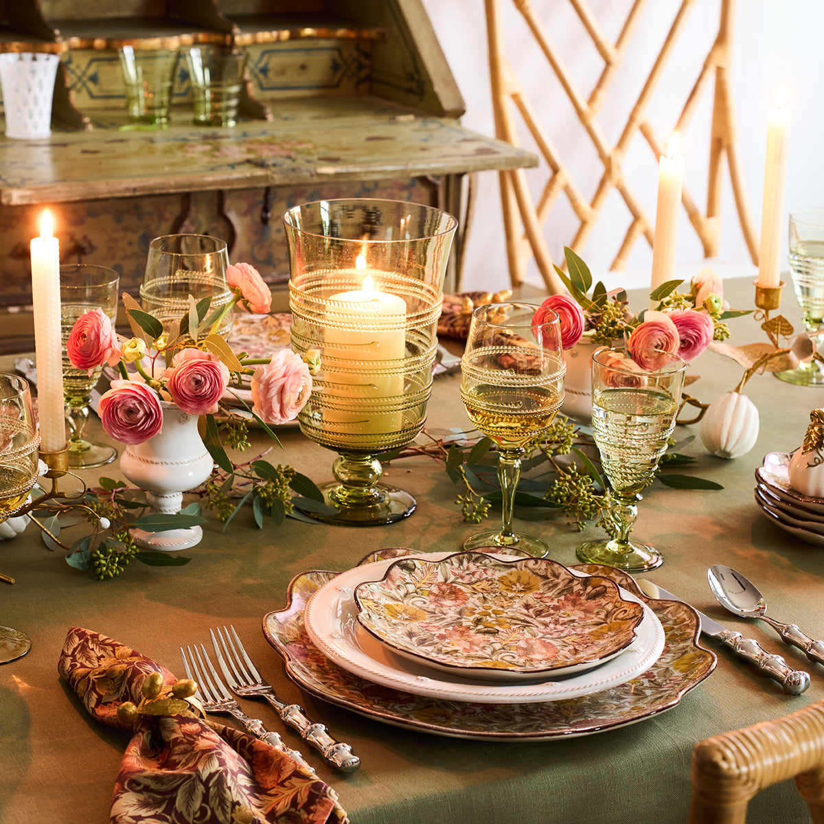 Styled with the Ella Drinkware Collection by Juliska, this elegant table features floral plates, artisan glasses, candles, and pink flower arrangements atop a green tablecloth, creating a warm and inviting atmosphere.