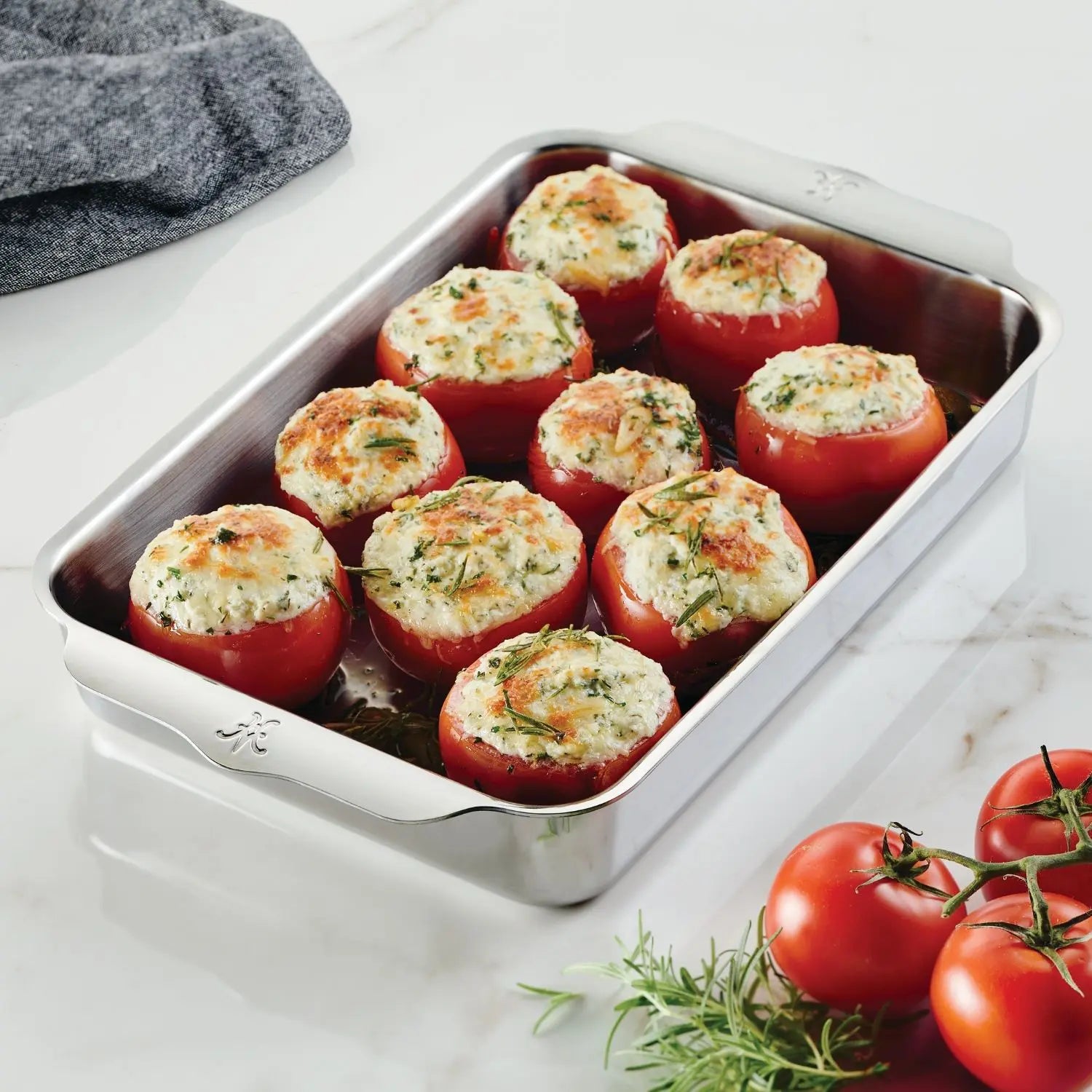The Hestan Provisions OvenBond Tri-ply Rectangular Baker, shown on a white countertop with fresh tomatoes and a gray cloth, holds nine baked stuffed tomatoes topped with herbs—ideal for holiday entertaining.