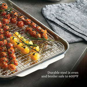 The Hestan Provisions OvenBond Tri-ply 5-Piece Set’s stainless steel pan with rack holds roasted tomatoes, with folded gray towels beside it. Warp-resistant, it's safe for oven and broiler use up to 600°F.