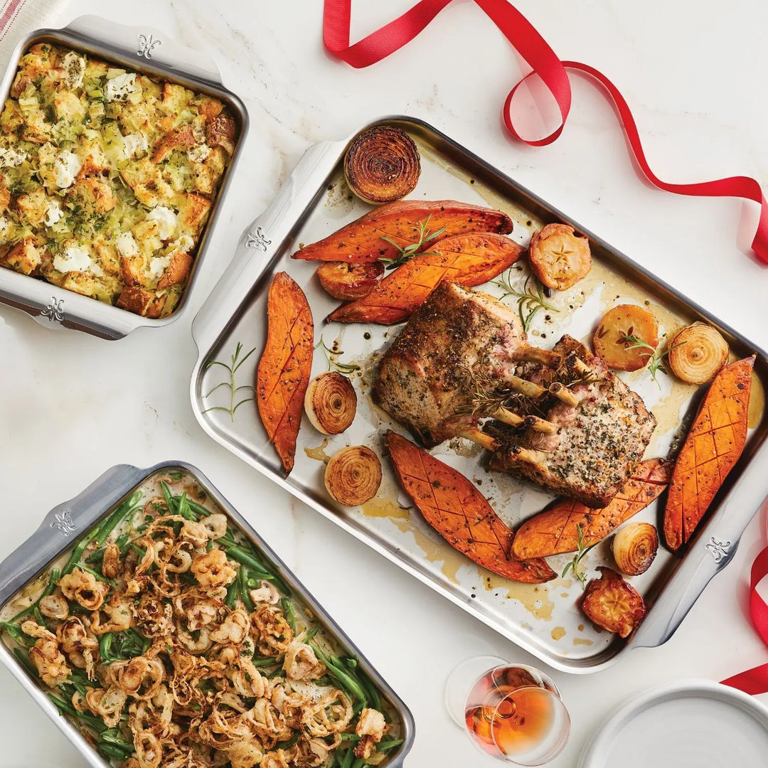 A roasted rack of pork with sweet potatoes, onions, green bean casserole, and herbed stuffing is artfully arranged on a Hestan Provisions OvenBond Tri-ply 5-Piece Set pan, set atop a white table adorned with a red ribbon.