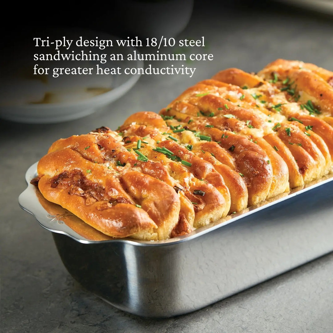 A golden-brown pull-apart bread loaf rests in a Hestan Provisions OvenBond Tri-ply Stainless Steel Loaf Pan, its superior heat conductivity ensuring even baking and perfectly sprinkled herbs on top.