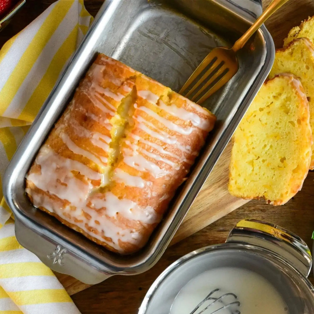 A loaf of glazed lemon bread sits on a wooden board with slices, a fork, and icing nearby—demonstrating how the Hestan Provisions OvenBond Tri-ply Stainless Steel Loaf Pan ensures even baking with superior heat conductivity.