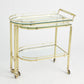 The Gallery Folding Bar Shiny Brass by Global Views features two glass shelves, curved edges, solid brass handles, and four wheels for mobility.