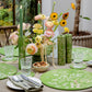 A dining table set with green floral placemats, matching napkins, glassware, candles, mixed nuts, and a vase of yellow and pink flowers features elegant Addison Ross Chinoiserie Salt and Pepper Mills, with a garden view outside.