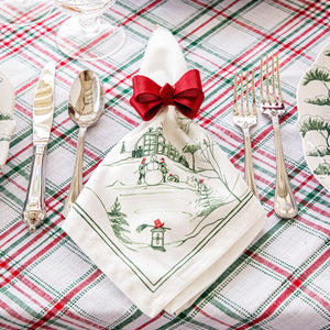 A holiday table is set with silverware and Juliska Country Estate Winter Frolic Evergreen Napkins, featuring a winter scene and red bow, atop a red, green, and white plaid tablecloth.