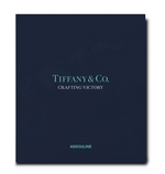 A navy blue book cover titled "Tiffany & Co.: Crafting Victory" in light blue text highlights the artistry of Tiffany hollowware, while the brand name "Assouline" is elegantly printed at the bottom, celebrating silversmithing excellence.