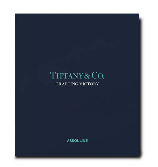 A navy blue book cover titled "Tiffany & Co.: Crafting Victory" in light blue text highlights the artistry of Tiffany hollowware, while the brand name "Assouline" is elegantly printed at the bottom, celebrating silversmithing excellence.